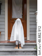 A child dressed as a ghost stands on a porch stoop, looking out towards the street, ready to trick-or-treat. Стоковое AI-изображение, AI-художник Данила Большаков / Фотобанк Лори