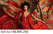 A woman in a red dress with beaded embellishments poses on a floral patterned rug. She is wearing a plunging neckline and her hair is styled in loose curls. Стоковое AI-изображение, AI-художник Данила Большаков / Фотобанк Лори