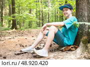 12-летний мальчик сидит на земле, прислонившись спиной к дереву в лесу. Стоковое фото, фотограф Яна Проничева / Фотобанк Лори