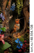 A Rufous hummingbird with a long, curved beak perches inside a hollowed-out tree branch, surrounded by vibrant blue flowers and greenery. Стоковое AI-изображение, AI-художник Данила Большаков / Фотобанк Лори