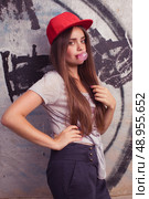 trendy beautiful long haired model posing on graffiti background. Blow bubblegum and show thumb up. red cap. grey t-shirt. Стоковое фото, агентство Ingram Publishing / Фотобанк Лори