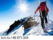 Mountaineer walking up along a steep snowy ridge with the skis in the backpack. In background a dramatic sky with a shiny bright sun. Concepts: adventure, achievement, courage, determination, self-realization, dangerous activity, extreme sport, winter leisure. Стоковое фото, агентство Ingram Publishing / Фотобанк Лори