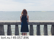 A young woman wearing a blue dress is standing by a wall with concrete balustrades on a promenade and is admiring the seaside on a sunny day. Стоковое фото, агентство Ingram Publishing / Фотобанк Лори