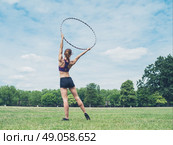 A fit and athletic young woman is standing on the grass in a park with a hula hoop raised above her head. Стоковое фото, агентство Ingram Publishing / Фотобанк Лори