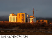 Вечерний вид на стройплощадку. Строительство многоквартирных жилых домов. Возведение нового комплекса разноэтажных зданий. Фасады освещены солнцем на закате. Башенный кран на строительной площадке. Жилищное строительство на Дальнем Востоке России. Город Магадан, Магаданская область, Россия. Стоковое фото, фотограф Андрей Степанов / Фотобанк Лори