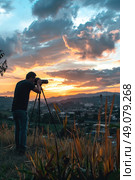 A photographer captures a stunning sunset over a cityscape, using a tripod to steady their camera. The golden hour light paints the clouds in hues of orange and pink, while the city lights twinkle below. Стоковое AI-изображение, AI-художник Данила Большаков / Фотобанк Лори