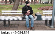 Pensive hipster drinking coffee sitting on a bench in the urban park. Handsome man wearing black coat eating street food, hot dog with paper cup in hand in the city. Stylish bearded middle-aged male. Стоковое видео, видеограф Марина Демидюк / Фотобанк Лори