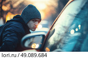 A young man wearing a beanie looks suspiciously at the camera while standing near a car, in a city environment at sunset. Стоковое AI-изображение, AI-художник Катерина Бондарец / Фотобанк Лори