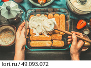 Tiramisu making.  Dish with italian savoiardi cookies and tiramisu ingredients on rustic kitchen table, top view. Woman hands holding a spoon and smeared cream on cookies.  Italian life style. Стоковое фото, агентство Ingram Publishing / Фотобанк Лори