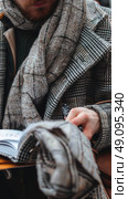 A close-up shot of a mans hand holding a pen over an open notebook. He is wearing a warm, checkered coat and a scarf. The photo is taken from a low angle, focusing on the mans hand and the notebook. Стоковое AI-изображение, AI-художник Данила Большаков / Фотобанк Лори