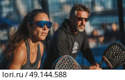 Focused woman and man playing padel outdoors, intense expression, teamwork, dynamic light and sporty mood on court with spectators. Стоковое AI-изображение, AI-художник Катерина Бондарец / Фотобанк Лори