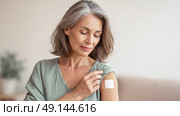 55-летняя женщина аккуратно прикрепляет пластырь гормональной терапии к плечу. Медицинская помощь, заместительная гормональная терапия в период менопаузы. Стоковое AI-изображение, AI-художник Катерина Бондарец / Фотобанк Лори