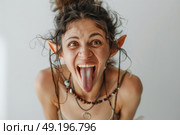 A woman with elf ears sticks her tongue out playfully, with her hair tied up and various beaded necklaces, standing against plain light background. Стоковое AI-изображение, AI-художник Данила Большаков / Фотобанк Лори