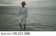 A young man in light white clothes and a hat walks barefoot in the rain on the water of the sea. Stormy evening clouds are in the background. Maldives. Стоковое видео, видеограф Aleksey Kidisyuk / Фотобанк Лори