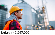 Worker in safety gear observing grain silos during twilight at an agricultural site with soft lighting effects. Стоковое AI-изображение, AI-художник Сергей Толмачёв / Фотобанк Лори