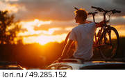 Sunset bike adventure at a scenic overlook with a young man preparing for a ride. Стоковое AI-изображение, AI-художник Сергей Толмачёв / Фотобанк Лори