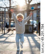 Blonde child in sweater joyfully plays with ball outdoors in sunlit urban setting. Стоковое AI-изображение, AI-художник Olga Didraga / Фотобанк Лори