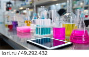Brightly colored chemicals in laboratory glassware on a stainless steel table with digital device in modern research facility. Стоковое AI-изображение, AI-художник Сергей Толмачёв / Фотобанк Лори