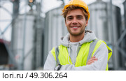 Young construction worker smiles confidently at a construction site with silos under overcast sky. Стоковое AI-изображение, AI-художник Сергей Толмачёв / Фотобанк Лори