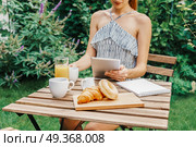 Young And Attractive Woman Having Morning Breakfast In Green Garden With French Croissant, Donuts, Coffee Cup, Orange Juice, Tablet and Notes Book On Wooden Table. Стоковое фото, агентство Ingram Publishing / Фотобанк Лори