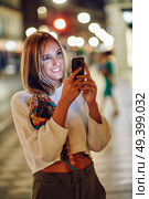 Blonde woman taking photograph with smartphone at night in the street. Defocused city lights at the background. Pretty girl with pigtail hairstyle at night smiling. Стоковое фото, агентство Ingram Publishing / Фотобанк Лори