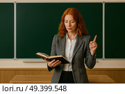 Женщина-преподаватель в деловом костюме стоит перед классной доской. Она держит открытую книгу в одной руке и подняла указательный палец другой, как будто объясняет что-то ученикам. Фон — зелёная школьная доска и часть деревянной панели. Освещение мягкое, интерьер типичного класса. Стоковое AI-изображение, AI-художник Sergey Starostin / Фотобанк Лори