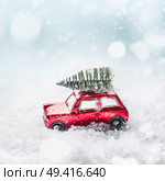 Christmas tree on red toy retro car in snow, through snowy winter wonder land with snowfall and bokeh. Creative Christmas holiday concept. Copy space for your greeting and design. . Стоковое фото, агентство Ingram Publishing / Фотобанк Лори