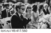 Man taking photos at outdoor wedding ceremony surrounded by guests in elegant attire during sunny day. Стоковое AI-изображение, AI-художник Сергей Толмачёв / Фотобанк Лори