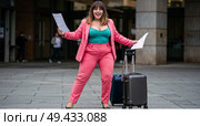Женщина plus-size в розовом брючном костюме взволнованно держит бумаги с чемоданом на открытом воздухе размытый фон. Стоковое AI-изображение, AI-художник Наталья Харинова / Фотобанк Лори