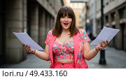 Женщина plus-size в розовом брючном костюме взволнованно держит бумаги с чемоданом на открытом воздухе размытый фон. Стоковое AI-изображение, AI-художник Наталья Харинова / Фотобанк Лори