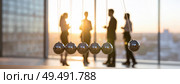 Business professionals in office silhouetted against sunrise with Newton’s cradle in foreground symbolizing corporate momentum, decision-making and teamwork. Стоковое AI-изображение, AI-художник Igor Tichonow / Фотобанк Лори