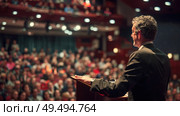 A man stands confidently at a podium, speaking to an attentive crowd of people gathered in front of him. Стоковое AI-изображение, AI-художник Данила Большаков / Фотобанк Лори