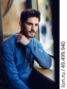 Thoughtful man with blue sweater with lost look near a window in a modern pub. Bearded guy with modern hairstyle. Life style concept.. Pensive man with blue sweater with lost look near a window. Стоковое фото, агентство Ingram Publishing / Фотобанк Лори