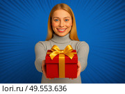 Молодая улыбающаяся блондинка с ярко-голубыми глазами в светло-сером свитере с высоким воротом. Она протягивает вперёд красную коробку с золотой подарочной лентой, создавая эффект вовлечённости и визуального контакта с аудиторией. Фон — насыщенно-синий с радиальными полосами в стиле комикса (поп-арт), создающими ощущение динамики и привлечения внимания. Композиция оформлена в горизонтальной ориентации 16:9, идеально подходящей для баннеров, обложек, слайдов или рекламных вставок в маркетплейсах и соцсетях. Свет ровный, студийный. Картинка выглядит как коммерческий шаблон, созданный для маркетинговых целей: продвижение акций, подарков, распродаж, оформления карточек товара. Стоковое AI-изображение, AI-художник Sergey Starostin / Фотобанк Лори