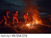 A group of firefighters in full gear standing around a fire, assessing the situation and preparing to extinguish the flames. Стоковое AI-изображение, AI-художник Данила Большаков / Фотобанк Лори