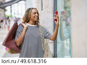 Young black woman photographing a store window in a shopping street with her smartphone. Female with african braids.. Young black woman photographing a store window in a shopping street with her smartphone. Стоковое фото, агентство Ingram Publishing / Фотобанк Лори