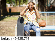 Young smiling African woman with skateboard. Mixed-race girl relaxing after riding skateboard listening to the music outdoors.. African woman with skateboard relaxing after riding skateboard listening to the music. Стоковое фото, агентство Ingram Publishing / Фотобанк Лори