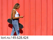 Young black girl with coloured braids walking on an urban red background while consulting her smartphone.. Black girl walking on an urban red background while consulting her smartphone. Стоковое фото, агентство Ingram Publishing / Фотобанк Лори