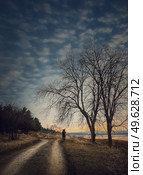 Lonely nomad person walking a country road. Moody and calm evening scene. Wanderer silhouette on trail. Cold season idyllic rural landscape with leafless trees and dry grass. Стоковое фото, агентство Ingram Publishing / Фотобанк Лори