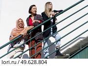Low angle of trendy young multiethnic female best friends, on stylish outfit and hijab, leaning on metal railing while standing on staircase and looking away in city on sunny day. Content young diverse ladies looking away while standing on stairs in city. Стоковое фото, агентство Ingram Publishing / Фотобанк Лори
