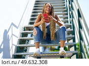 Black girl with coloured braids, sitting on some steps with a smartphone and a skateboard.. Black girl with coloured braids, sitting with a smartphone and a skateboard. Стоковое фото, агентство Ingram Publishing / Фотобанк Лори