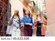 Positive multiracial female friends with photo camera standing on street against building and looking up happily. Diverse stylish women amused near building. Стоковое фото, агентство Ingram Publishing / Фотобанк Лори
