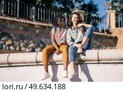 Low angle of cheerful multiethnic female best friends cuddling and sitting on concrete border in street while looking away. Positive diverse women sitting on fence in city. Стоковое фото, агентство Ingram Publishing / Фотобанк Лори