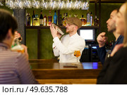 Glad bearded man, in white shirt and cap, smiling and mixing cocktail ingredients in shaker, while standing behind counter near male and female customers in bar. Positive barkeeper preparing cocktail for clients in a pub. Стоковое фото, агентство Ingram Publishing / Фотобанк Лори
