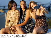 Smiling charming multiracial girlfriends sitting together on stone bench and taking self portrait on cellphone while enjoying pleasant time. Cheerful diverse girlfriends taking selfie on smartphone in sunny day. Стоковое фото, агентство Ingram Publishing / Фотобанк Лори