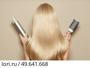Back view of woman with long beautiful blond hair isolated on beige background holds hairdressing accessories. Dyeing and hair care. Shiny smooth blonde hair. Стоковое фото, агентство Ingram Publishing / Фотобанк Лори