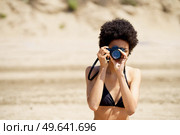 African American female photographer in swimwear taking picture on photo camera while standing on sandy shore during trip in tropical resort. Black woman in bikini taking photo on beach. Стоковое фото, агентство Ingram Publishing / Фотобанк Лори