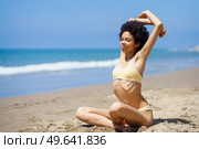 Full body of young positive African American female millennial with curly dark hair in trendy bikini, stretching arms while warming up on sandy coast against cloudless blue sky and sea. Smiling black lady stretching arms on sandy beach during summer vacation. Стоковое фото, агентство Ingram Publishing / Фотобанк Лори