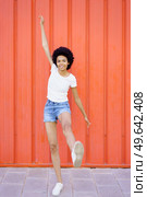 Full body of optimistic African American female in casual clothes looking at camera with raised arm while standing on one leg on red background. Glad black woman with raised leg. Стоковое фото, агентство Ingram Publishing / Фотобанк Лори