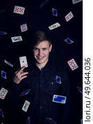 Successful young man holds up a victory lucky card. Gambler play luck games and win, isolated on dark background. Gambling jackpot winner with an ace of hearts under a rain of playing cards. Стоковое фото, агентство Ingram Publishing / Фотобанк Лори